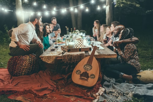 Friends gathered outdoors around a table full of food.There are hanging white outdoor lights and blankets on the ground with a guitar.