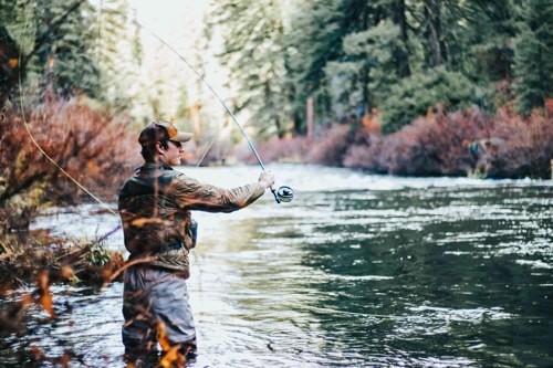Middle-aged man in camo shirt and hat and blue jeans fly fishing in a river.