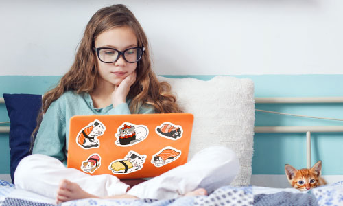 Teen sitting on her bed with a laptop that has kawaii animal stickers on the case | Stickers.com